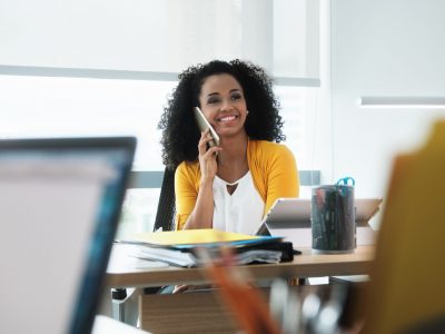 Young business woman working in modern office. Black businesswoman talking and smiling on mobile telephone. Busy African American employee using smartphone at work